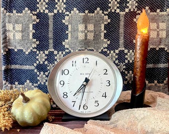 May include: A vintage silver-tone Westclox Big Ben clock with a white face and black numbers. A small, light green pumpkin and a brown candle sit nearby. The background features a blue and white patterned fabric.
