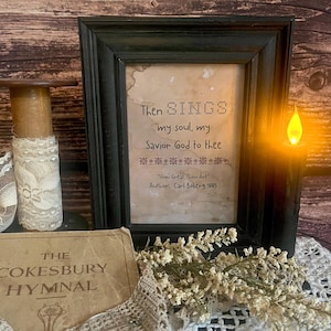 May include: A framed print with a black frame and a white background. The print features a cross-stitch design with the text "Then SINGS my soul, my Savior God to thee." The text "How Great Thou Art" and "Author: Carl Boberg 1885" are also included.