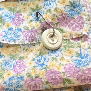 May include: A white button with a metal pin attached to a piece of fabric with a floral pattern in shades of blue, yellow, and purple.