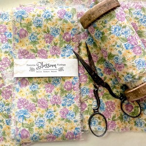 May include: A close-up of a floral fabric with a light yellow background, featuring pink and blue flowers. The fabric is rolled up on a wooden spool and there are vintage scissors nearby. The fabric has a frayed edge and a label that reads "Prairie Blossom Vintage Revive Restore Relove".