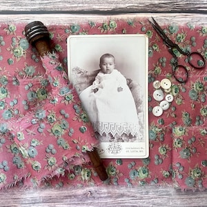 May include: A vintage photograph of a baby in a white dress with lace trim, resting on a floral patterned fabric. The image is accompanied by a wooden spool, antique scissors, and several white buttons. The fabric has a pink background with blue and green floral accents.