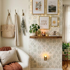 May include: A room corner featuring a white armchair with a rust-colored throw blanket, a woven tote bag, a wall-mounted wooden shelf with a potted plant, a candle, a mug, and a book, plus four framed wall art prints with the text STAY COZY.