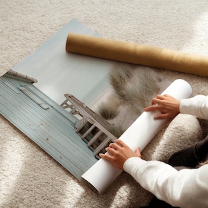 May include: A large rectangular photographic print featuring a coastal scene with a weathered light blue wooden structure, a sandy path, and dune grass. The print is partially rolled, with a brown cardboard shipping tube lying nearby.