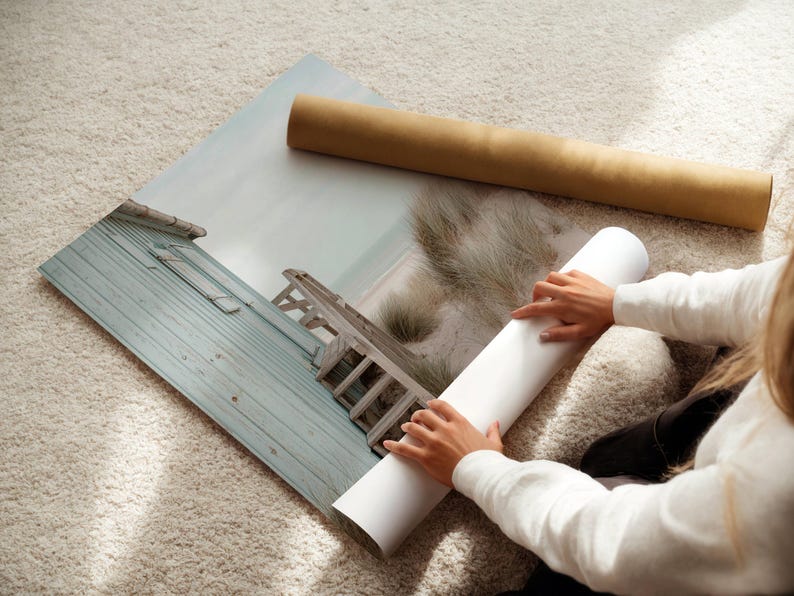 May include: A large rectangular photographic print featuring a coastal scene with a weathered light blue wooden structure, a sandy path, and dune grass. The print is partially rolled, with a brown cardboard shipping tube lying nearby.
