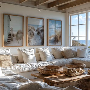 May include: Three framed wall art prints depicting coastal beach scenes with sand dunes, tall grasses, and wooden structures, displayed above a large white sectional sofa with textured throw pillows and a wooden coffee table with woven baskets.