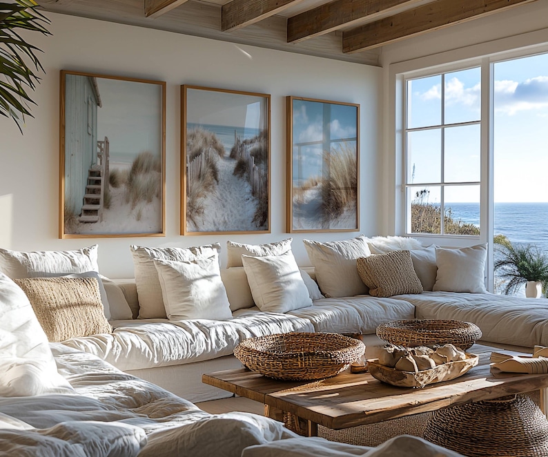 May include: Three framed wall art prints depicting coastal beach scenes with sand dunes, tall grasses, and wooden structures, displayed above a large white sectional sofa with textured throw pillows and a wooden coffee table with woven baskets.