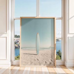 May include: A framed vertical wall art print featuring a light blue surfboard standing upright in the center of a sandy beach. The background displays a calm ocean horizon under a clear sky, all enclosed within a simple light-toned wooden frame.