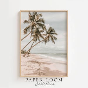 Puede incluir: Una impresión de arte mural enmarcada vertical que muestra una playa de arena con palmeras inclinadas hacia el océano bajo un cielo nublado. El texto PAPER LOOM Collection está impreso en negro en la parte inferior central de la imagen.