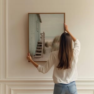 May include: A rectangular wall art print in a thin brown frame featuring a coastal scene with a light green wooden beach hut, a set of wooden stairs leading to the sand, and tall beach grass under a soft, overcast sky on a white wall.