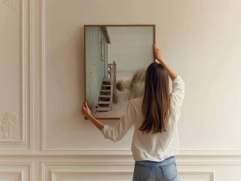 May include: A rectangular wall art print in a thin brown frame featuring a coastal scene with a light green wooden beach hut, a set of wooden stairs leading to the sand, and tall beach grass under a soft, overcast sky on a white wall.