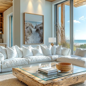 May include: A large white sectional sofa with multiple cushions, a low square coffee table with a thick natural wood base and glass top, a woven bowl, and a framed wall print depicting a sandy beach path with wooden fencing and ocean views.