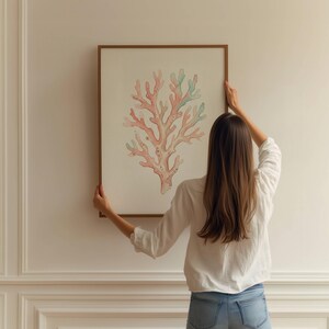 May include: A rectangular wall art piece featuring a detailed illustration of a branching coral structure in shades of soft pink and light green, encased in a thin wooden frame, displayed against a plain, off-white paneled wall surface.
