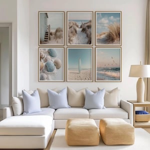 May include: A gallery wall featuring six framed coastal prints arranged in two rows. The images depict a beach house, sand dunes, tall grass, seashells, a surfboard on sand, and ocean waves with birds, all set in light wood frames above a sofa.