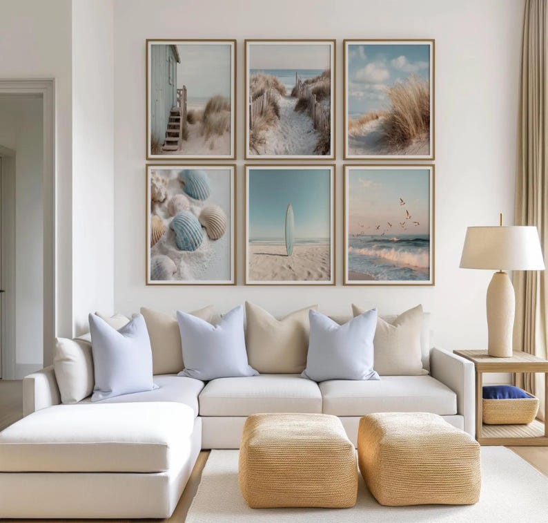 May include: A gallery wall featuring six framed coastal prints arranged in two rows. The images depict a beach house, sand dunes, tall grass, seashells, a surfboard on sand, and ocean waves with birds, all set in light wood frames above a sofa.