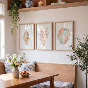 May include: A set of three framed wall art prints featuring watercolor illustrations of a spiral seashell, a branching coral, and a fan-shaped scallop shell in soft pastel shades of pink, blue, and peach, displayed in light wood frames.