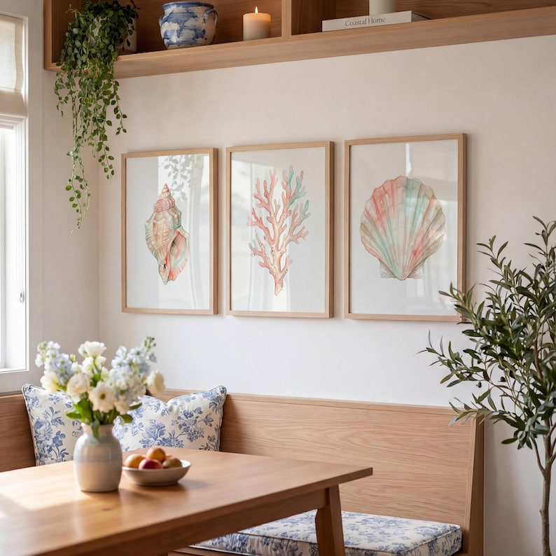 May include: A set of three framed wall art prints featuring watercolor illustrations of a spiral seashell, a branching coral, and a fan-shaped scallop shell in soft pastel shades of pink, blue, and peach, displayed in light wood frames.