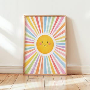 Op de afbeelding: Een rechthoekige wandkunstprint met een centrale gele zon met een eenvoudig zwart lijngezicht, inclusief twee ogen, een gebogen glimlach en roze wangen. De zon is omringd door stralende strepen in pastel regenboogkleuren, waaronder roze, oranje, geel, groen, blauw en paars.
