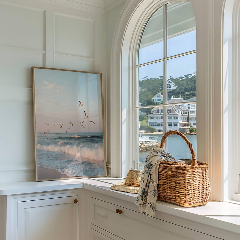 May include: A framed rectangular wall art print depicting a coastal scene with ocean waves and flying seagulls, positioned next to a woven wicker basket with a handle, a straw hat, and a patterned fabric draped over the basket's edge.