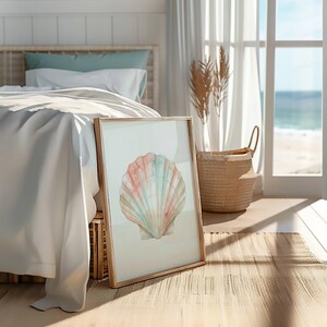 May include: A framed wall art print featuring a watercolor illustration of a single scallop shell in soft pastel shades of pink, peach, and light blue. The artwork is displayed in a light-toned wooden frame, resting on a woven floor surface.