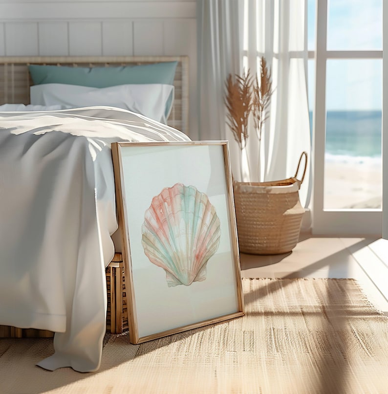 May include: A framed wall art print featuring a watercolor illustration of a single scallop shell in soft pastel shades of pink, peach, and light blue. The artwork is displayed in a light-toned wooden frame, resting on a woven floor surface.