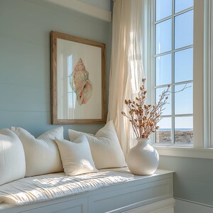 May include: A framed watercolor print of a pink and tan seashell hangs on a light blue wall above a window seat. Three off-white textured throw pillows sit on the bench next to a white ceramic vase filled with dried brown botanical branches.