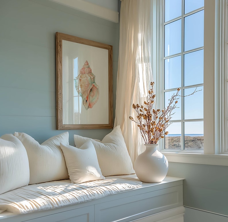 May include: A framed watercolor print of a pink and tan seashell hangs on a light blue wall above a window seat. Three off-white textured throw pillows sit on the bench next to a white ceramic vase filled with dried brown botanical branches.