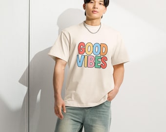 May include: A person wearing a cream-colored t-shirt with the words "GOOD VIBES" in colorful bubble letters. The person is also wearing light blue jeans and black and white sneakers. A silver chain necklace is visible.