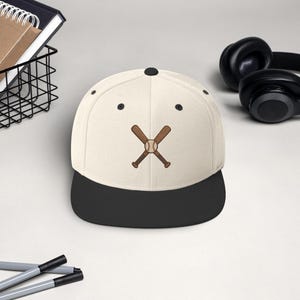 Op de afbeelding: Crèmekleurige baseballpet met een zwarte klep. De pet heeft een geborduurd ontwerp van gekruiste honkbalknuppels en een honkbal. Een stijlvol accessoire voor honkbalfans.