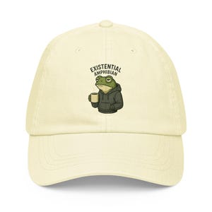 May include: Cream-colored baseball cap featuring an embroidered cartoon frog wearing a hoodie and holding a coffee mug. The text "EXISTENTIAL AMPHIBIAN" is arched above the frog. The cap has a curved brim and an adjustable strap.
