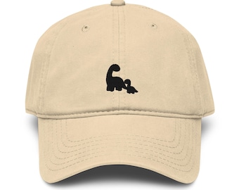 May include: A beige baseball cap with a curved brim. The cap features a black silhouette of a parent dinosaur and a baby dinosaur on the front. The cap has a classic six-panel design with a button on top.