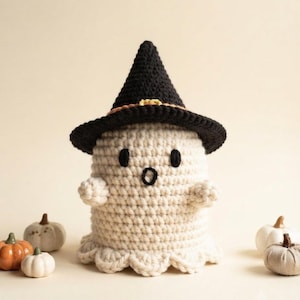 May include: A cream-colored crocheted ghost figure wearing a black pointed witch hat with an orange band and a small yellow buckle. The ghost has two black oval eyes and a small black circular mouth, with a scalloped bottom edge and small arms.