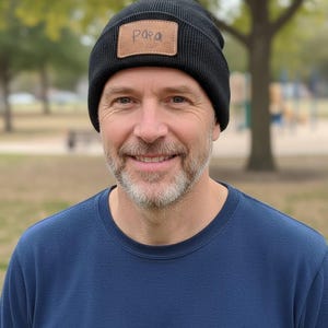 May include: Black knit beanie with a brown leather patch that reads "Papa". The beanie is worn by a person wearing a blue crew neck shirt. The background is a park with trees and grass.