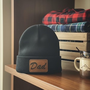 May include: A black beanie with a brown leather patch that reads "Dad". The hat is sitting on a wooden shelf, with folded plaid shirts and a mug with a mountain design in the background. A pair of glasses rests on the mug.