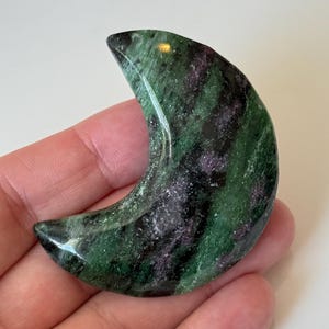 May include: A crescent moon-shaped gemstone with a polished surface. The stone features a mix of green and dark purple hues, creating a marbled effect. The gemstone is held in a hand.