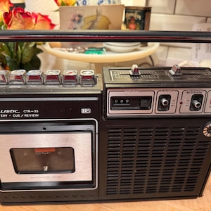 May include: A vintage black and silver Realistic CTR-33 cassette player with a handle. It features a cassette tape slot, buttons, and dials. The text "Realistic CTR-33 AC/Battery Cue/Review" is visible on the front.