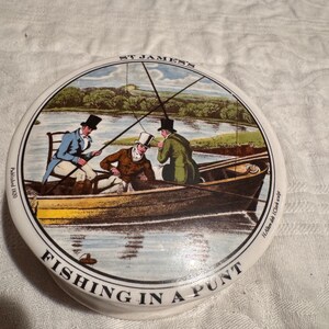 May include: A round, white ceramic dish with a detailed illustration of three men fishing in a punt. The image is framed by the text "ST JAMES'S" and "FISHING IN A PUNT". The illustration features a river scene with trees and a boat.