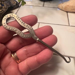 May include: A silver shoe horn with a loop handle. The handle is engraved with "THE SHOE MART CO." and "15TH ST., DENVER, COLO."