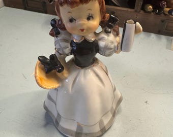 May include: A porcelain figurine of a girl wearing a yellow hat and a black and white dress. She is holding a book in one hand and a basket in the other. The figurine is painted in a realistic style and has a delicate, whimsical look.