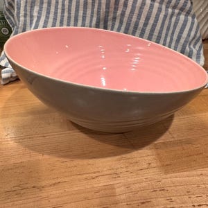 May include: A large, oval-shaped ceramic bowl with a light gray exterior and a pink interior. The bowl has a smooth, glossy finish and a slightly asymmetrical design. The bowl is sitting on a wooden surface.
