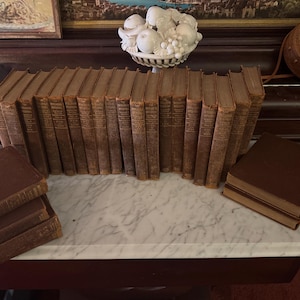 May include: A collection of antique books with brown leather covers. The books are arranged in a row, with some stacked on a marble surface. The spines show titles and are aged. A decorative fruit bowl and artwork are in the background.
