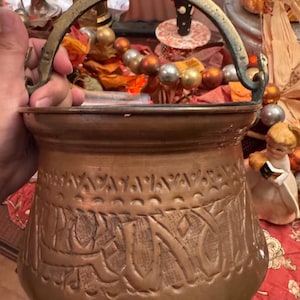 May include: A round copper pot featuring a swing handle and an embossed decorative band around the upper section. The metal surface displays a warm, metallic tone with visible signs of age and patina, including darkened areas on the handle.