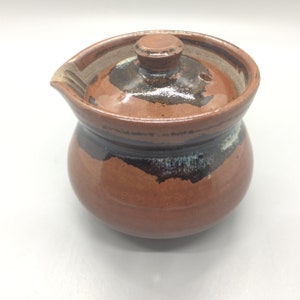 Puede incluir: Una tetera de cerámica hecha a mano con tapa. La tetera es de color marrón cálido con una banda negra alrededor del centro y un pico para verter. La tapa tiene un pequeño pomo y un agujero para que escape el vapor.