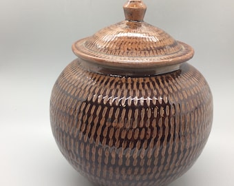 Op de afbeelding: Een bruine keramische pot met een deksel, met een getextureerd patroon. De pot heeft een rond lichaam en een licht uitlopende opening. Het deksel is gewelfd en heeft een kleine knop bovenop.