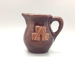 May include: A small brown ceramic creamer with a light brown geometric design of a house on the front.