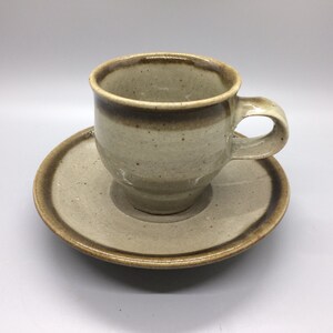 May include: A speckled beige ceramic coffee cup with a brown rim and a matching saucer. The cup has a rounded shape and a handle.