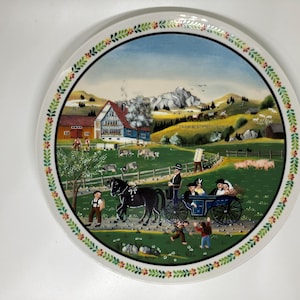 May include: A decorative ceramic plate featuring a pastoral scene. The plate's center depicts a horse-drawn carriage, houses, and mountains. The border has a floral design. The plate is white with a glossy finish.