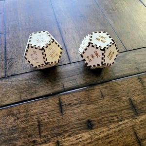 May include: Two wooden, multi-sided dice with laser-etched text. The dice are light brown with dark brown etched details. The text includes numbers and letters. The dice are on a dark brown wooden surface.