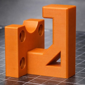 Puede incluir: Formas geométricas naranjas impresas en 3D. Las formas incluyen un ángulo recto, un bloque con una parte superior curva y un bloque con tres agujeros circulares. Los objetos tienen una superficie texturizada y están sobre un fondo gris.