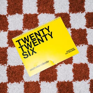 Könnte beinhalten: Ein gelber Kalender mit dem Text "TWENTY TWENTY SIX" in großen schwarzen Buchstaben. Der Kalender liegt auf einem braun-weiß karierten Teppich. Der Kalender trägt auch den Text "couch view calendar" und "PHOTOS BY ZAC WILLIAMS".
