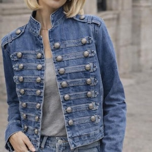 Könnte beinhalten: Blaue Jeansjacke mit militärisch inspiriertem Design. Die Jacke hat einen hohen Kragen, mehrere Reihen dekorativer silberner Knöpfe und eine taillierte Passform. Das Model trägt ein graues Shirt und blaue Jeans.